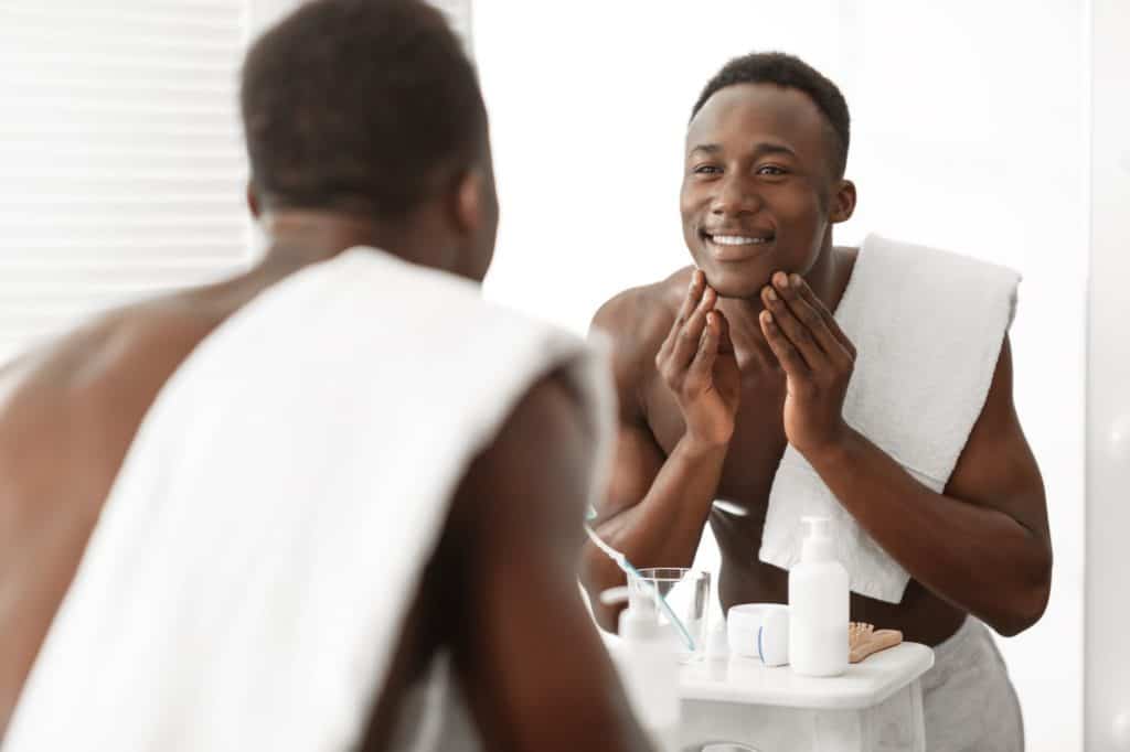 Black Man Touching Face Skin Standing Near Mirror In Bathroom